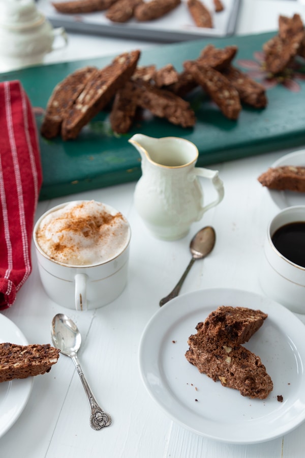 A white plate with chocolate biscotti on it and a green board with biscotti piled on it. Two cups of coffee in white cups and small silver spoons and a white pitcher of vegan cream and a red and white striped towel on a white wood table.