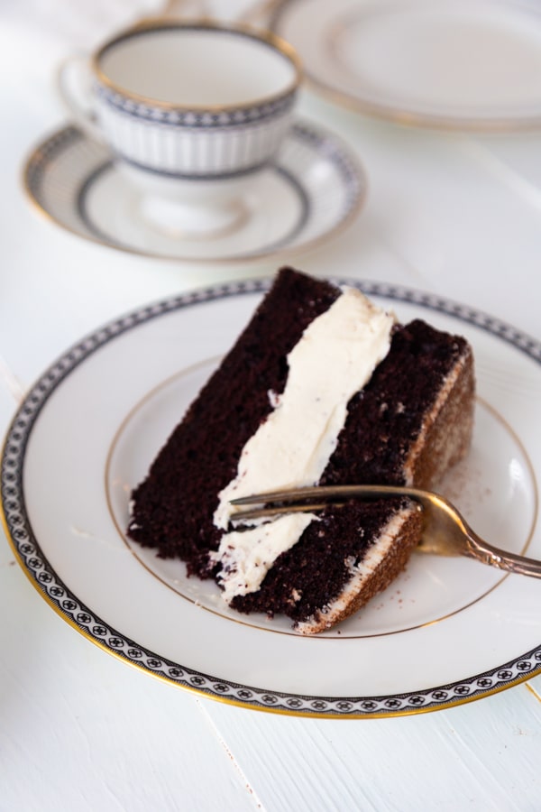 A slice of chocolate cake with vanilla frosting on a white plate with a silver fork cutting into it.