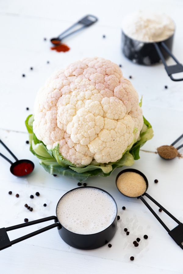 A whole cauliflower and assorted spices in black measuring cups and spoons on a white table.
