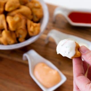 Veganosity's favorite recipe caulflower poppers being dipped in a vegan ranch sauce with a bowl of cauliflower poppers in the background with vegan ranch, vegan sriracha mayo, and vegan hot sauce in three separate white dishes on a wood board.