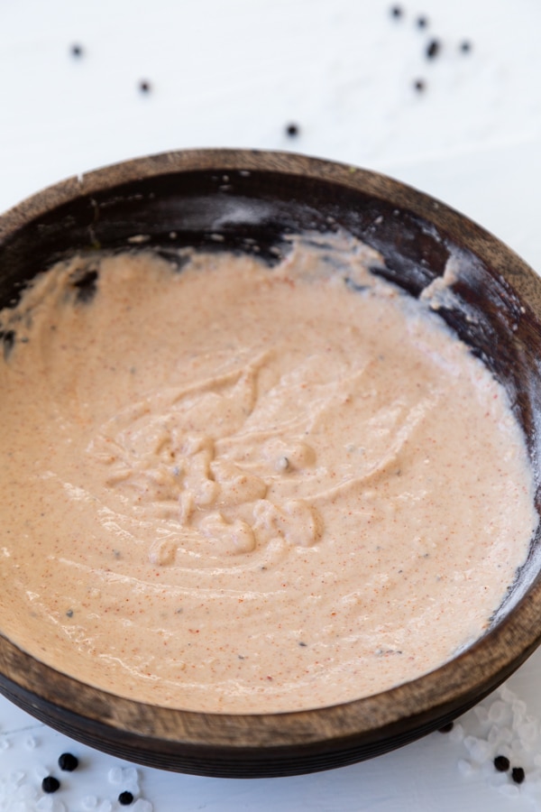 A wood bowl with a dipping batter on a white table with peppercorns scattered around it.