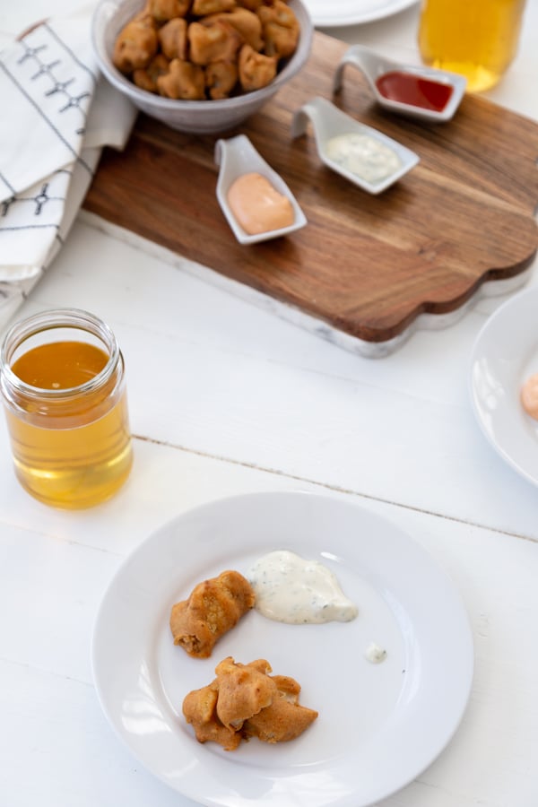 A white plate with 2 cauliflower poppers and white sauce and a glass of beer and a bowl of poppers and dipping sauces in the background.