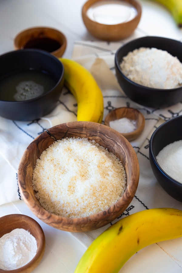 Wood bowls and black measuring cups with cake ingredients and two bananas on a white wood table.