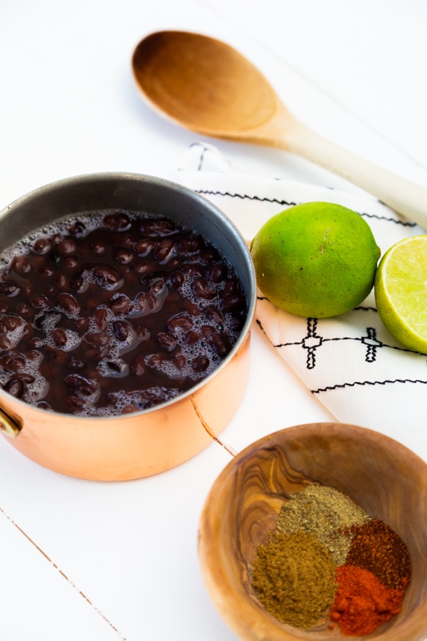 A copper pot with black beans and limes and a wood spoon next to the pot