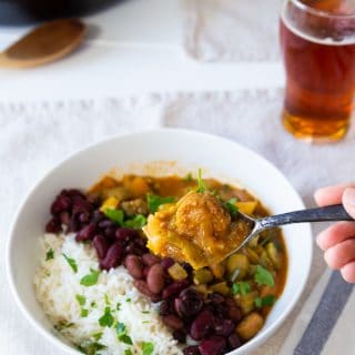 Gumbo in a white bowl with beans and rice with a hand holding a spoon of the stew, and a glass of beer and a skillet of gumbo in the background.