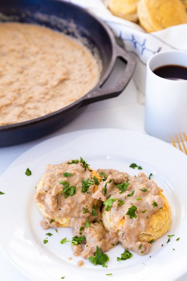 Biscuits and gravy on a white plate with a cup of coffee and a skillet of gravy in the background