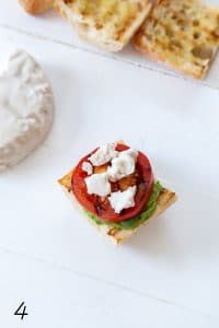 caprese sandwich with avocado pesto, grilled tomato, basil, and vegan mozzarella on a white board with a wheel of mozzarella
