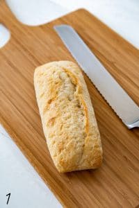 caprese sandwich bread on a wood cutting board with serrated knife on white wood