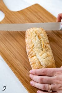 baguette being cut in half with serrated knife on a wood cutting board on top of white wood