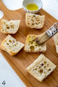 white wood board with wood cutting board on top with slices bread being brushed with olive oil