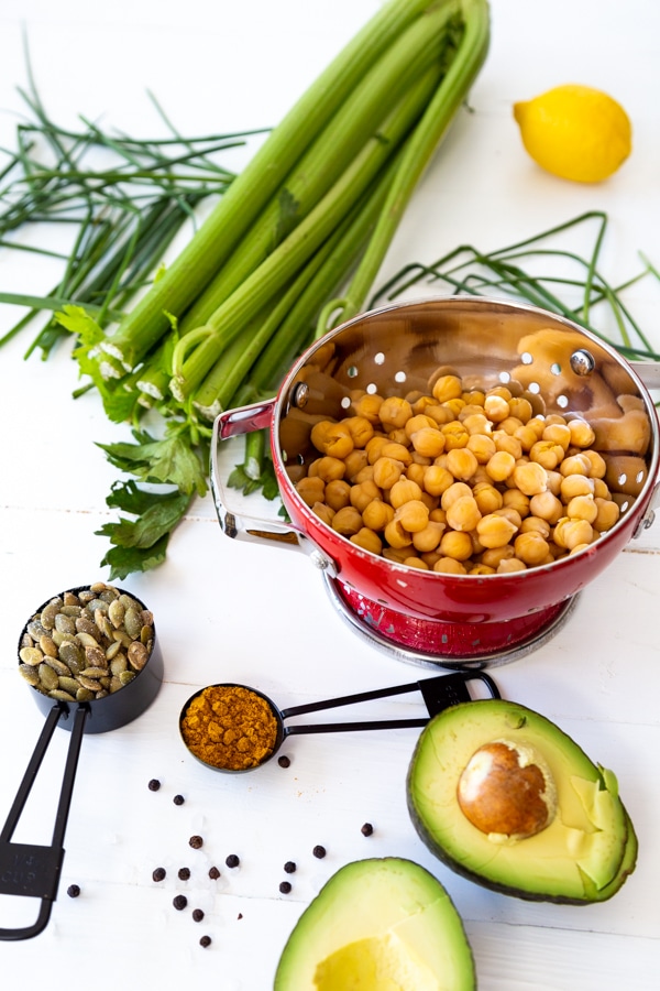 A red colander with chickpeas, black measuring spoons with spices, avocado, celery and chives on a white board.