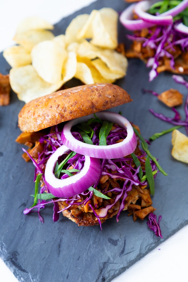 A pulled BBQ sandwich with onion and slaw and potato chips in the background.