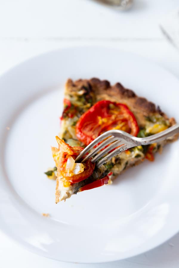 A slice of tomato tart on a white plate with a silver fork cutting into it.