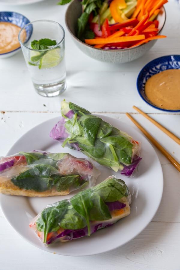 A white plate with three spring rolls and a blue bowl with peanut sauce, a glass of water, a bowl of veggies, and chopsticks on a white table. 
