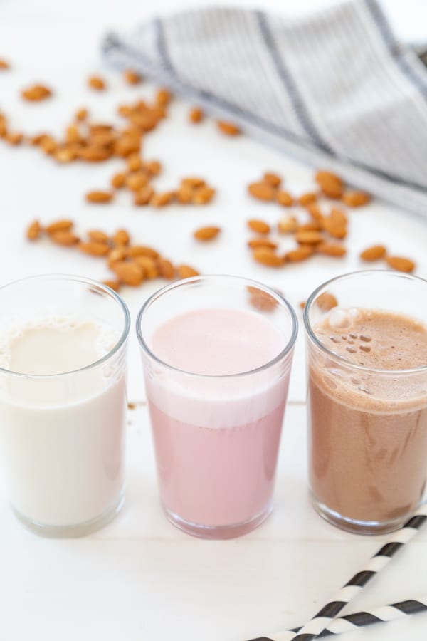 A glass of almond milk, strawberry milk, and chocolate milk in a row with chocolate chips and almonds scattered on a white table. 