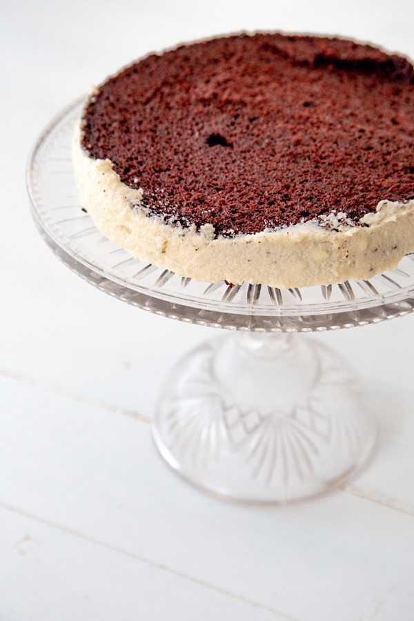 The bottom half of a chocolate cake on a glass cake stand with white frosting on the sides.