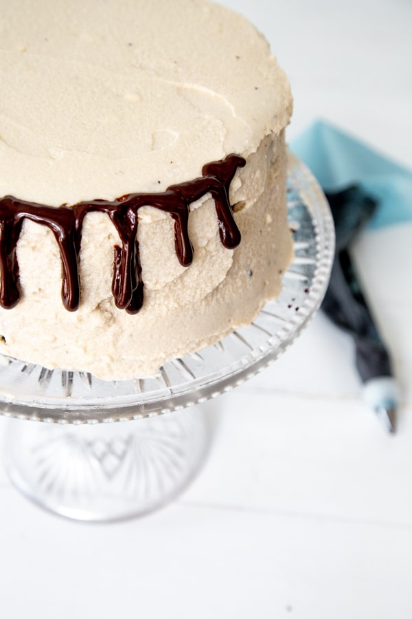A cake frosted with vanilla icing with drips of chocolate ganache down the sides and a blue pastry bag next to it.