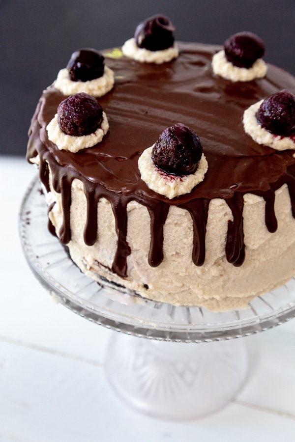 A cake with vanilla frosting and chocolate ganache on top and dripping down the sides with dollops of frosting and cherries on top of the dollops.