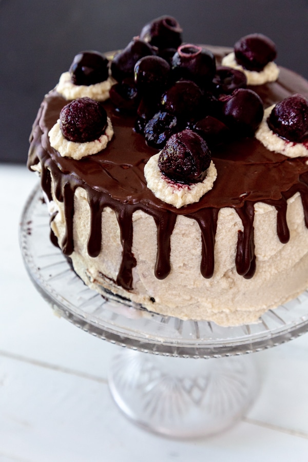A cake with vanilla frosting, chocolate ganache dripping down the sides, and cherries on top.