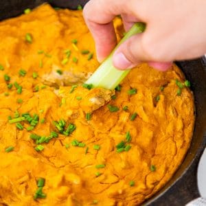 An iron skillet with buffalo chik'n dip and a hand dipping a stalk of celery in it.