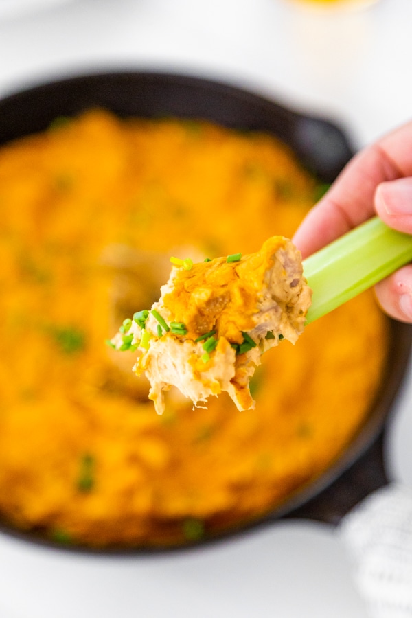 An iron skillet filled with buffalo chicken dip with a hand dipping a stick of celery in the center.
