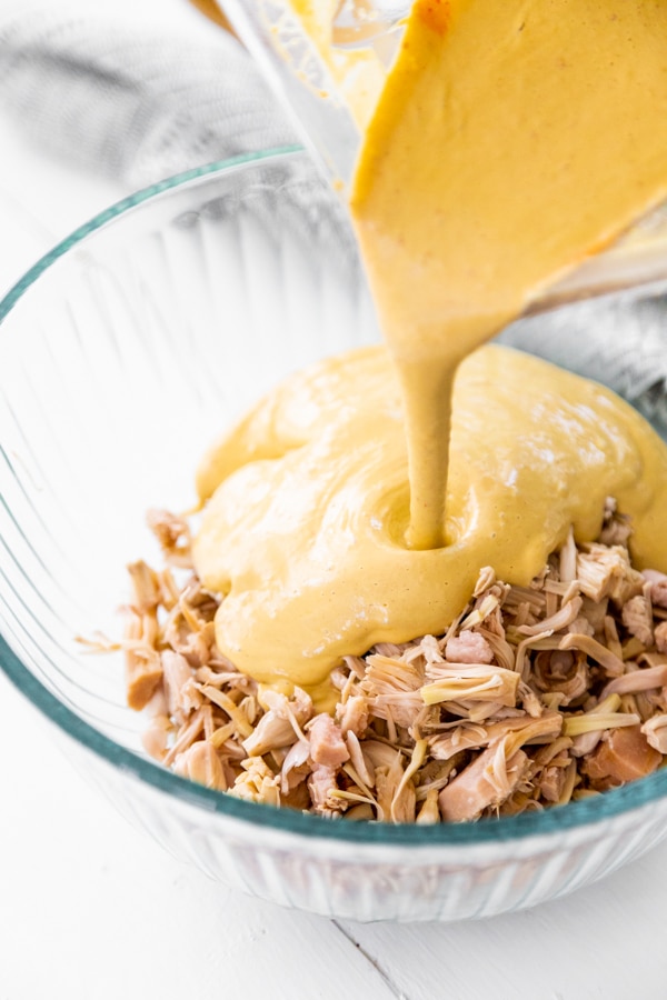 Cheese sauce being poured over shredded jackfruit in a glass bowl.