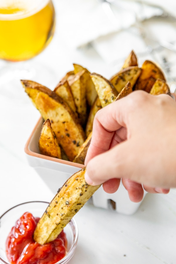 A hand dipping a home fry in a small dish of ketchup with a basket of fries in the background.