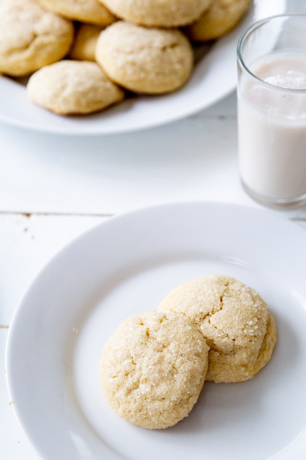 Two sugar cookies on a white plate with the platter of cookies and a glass of milk in the background.