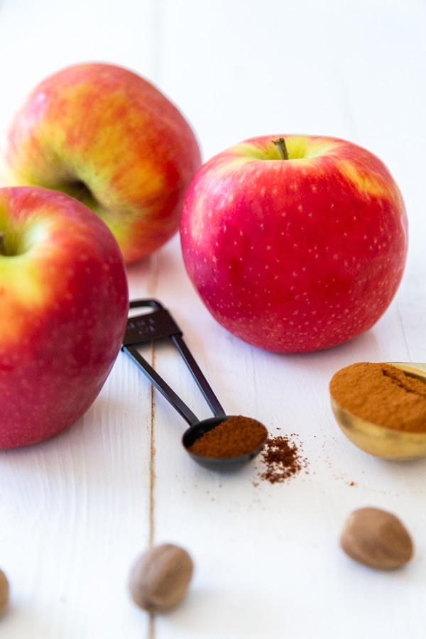 3 red apples and spices in measuring spoons and whole nutmeg on a white wood table.