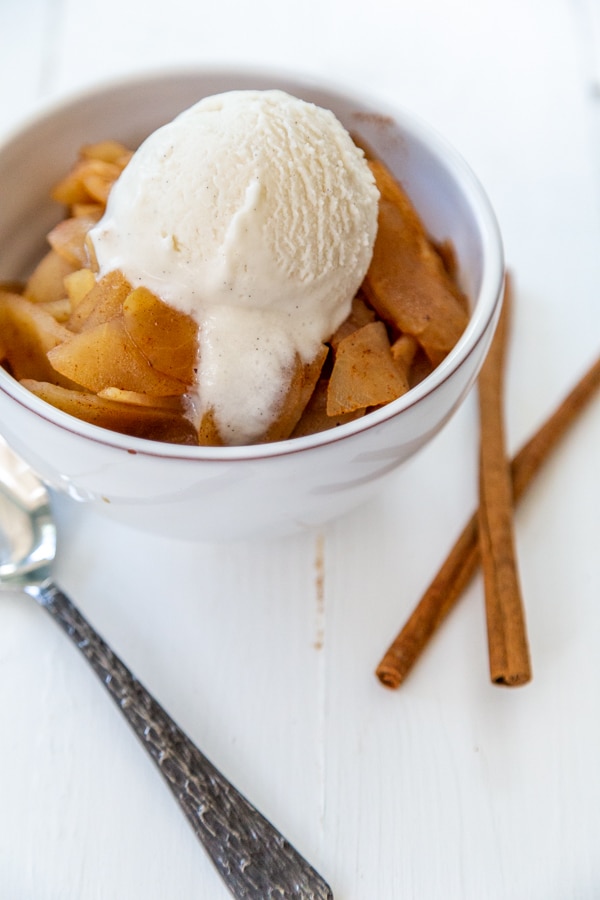 A white bowl filled with applesauce and ice cream with cinnamon sticks and a spoon next to it.