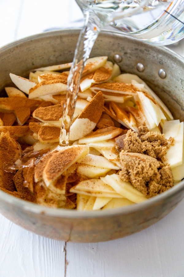 A copper pan with sliced apples and cinnamon and a pitcher of water being poured on to the apples.