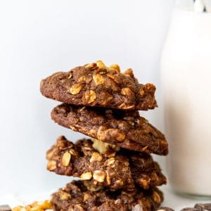 Four double chocolate chip cookies stacked with walnuts and a glass of milk with a straw in it
