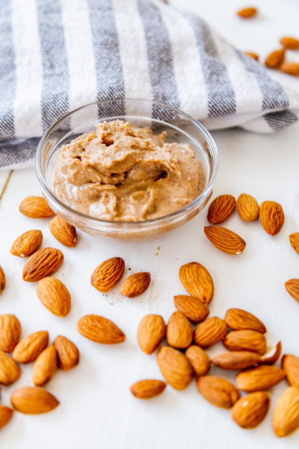 A glass dish filled with almond butter and almonds and a striped towel on a white wood table. 