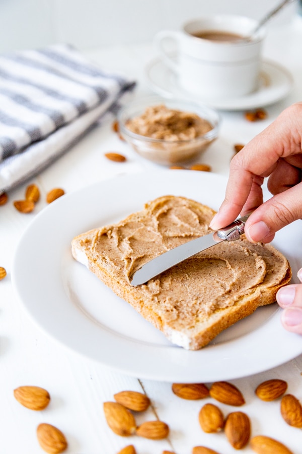 A hand spreading almond butter on a piece of toast on a white plate. 