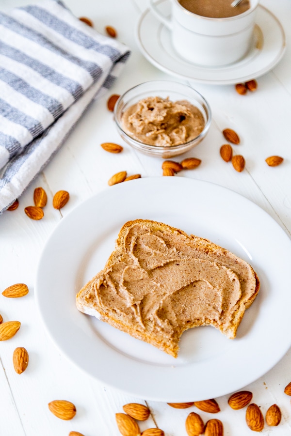 A piece of toast with almond butter with a bite taken out of it on a white plate with a cup of coffee and almonds spread around it. 
