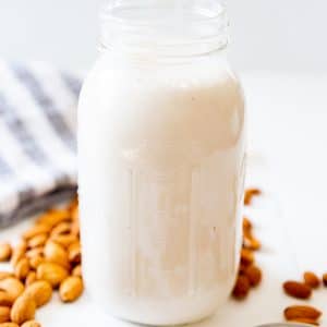 A jar of almond milk with almonds scattered next to it on a white wood table.