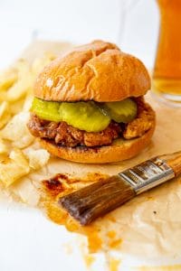 A spicy fried chicken sandwich on parchment paper with a pastry brush dripping spicy sauce next to it.