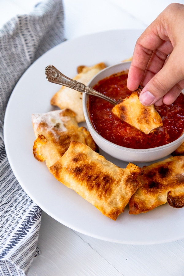 A white plate and bowl with pizza rolls and a hand dipping a roll into pizza sauce.