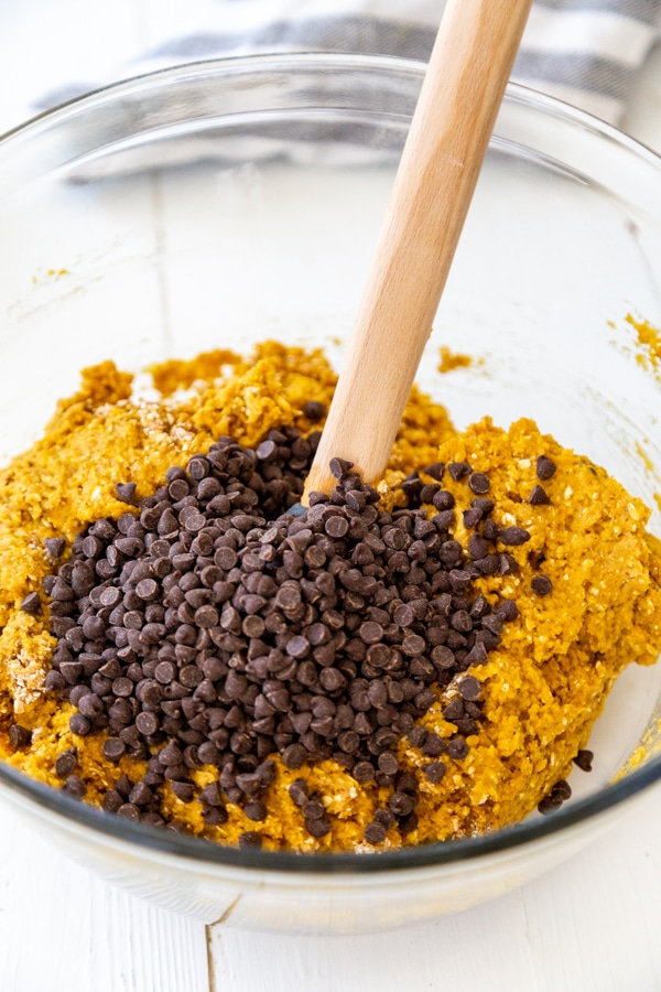 A glass bowl with pumpkin blondie batter and chocolate chips, with a wood spatula.