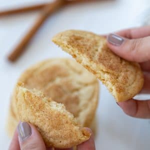 A hand breaking a cookie in half over a stack of cookies and cinnamon sticks.