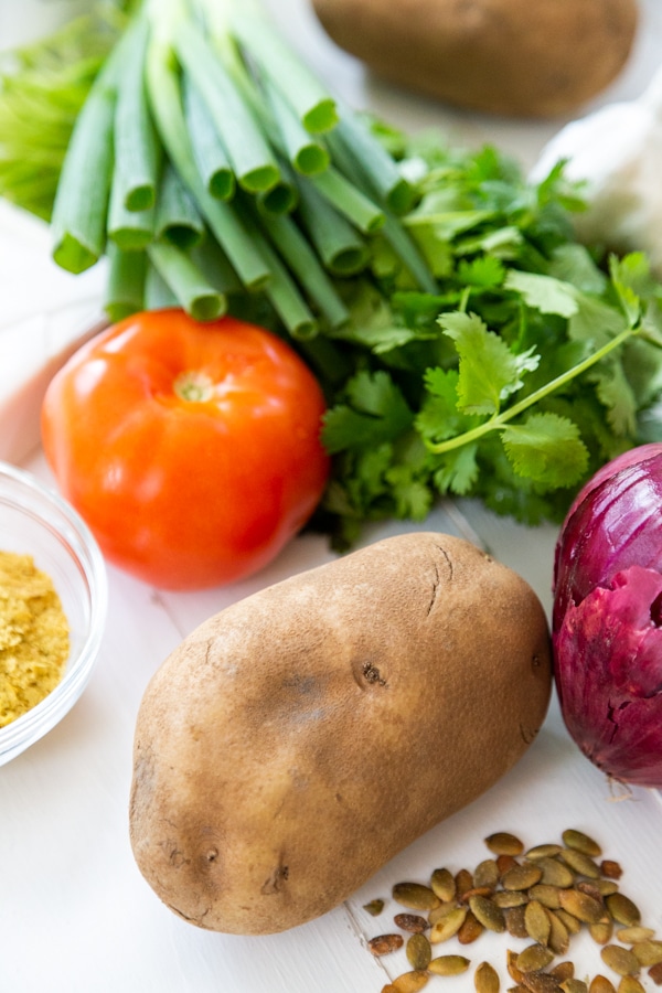 Potatoes, tomatoes, spices, pepitas, onion, and cilantro on a white wood table.