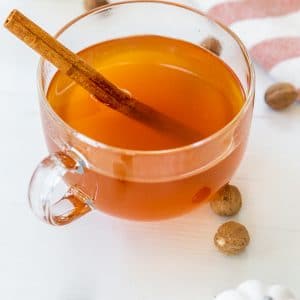 A clear glass mug of mulled apple cider with a cinnamon stick and white pumpkins, spices, and a towel on a white table.
