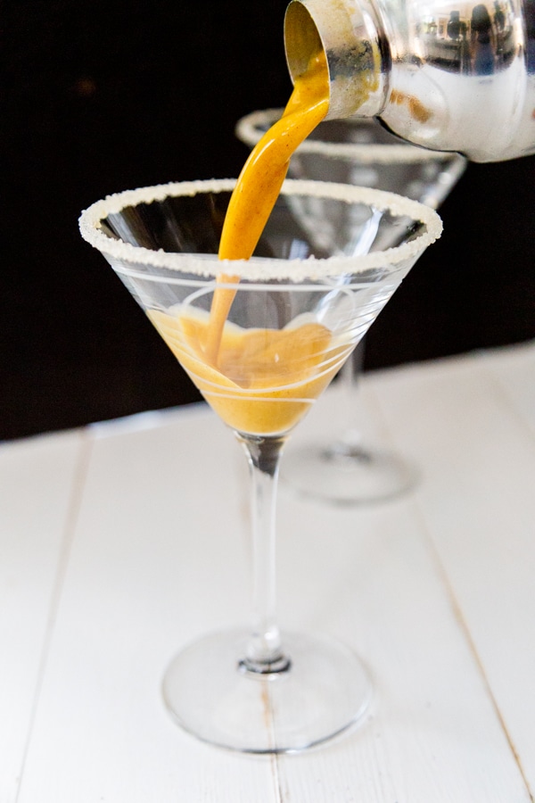 A hand pouring a shaker of pumpkin martini into a martini glass.
