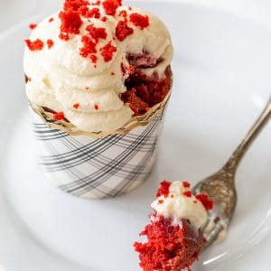 A red velvet cupcake with cream cheese frosting on a white plate with a fork full of the cake resting on the plate.