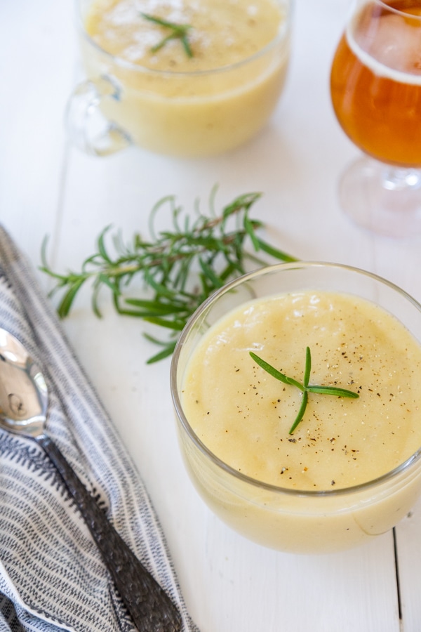 2 clear mugs of potato soup with black pepper and rosemary and a glass of beer next to the soup.