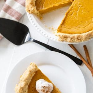 A slice of pumpkin pie with whipped topping on a white plate and the whole pie next to it.