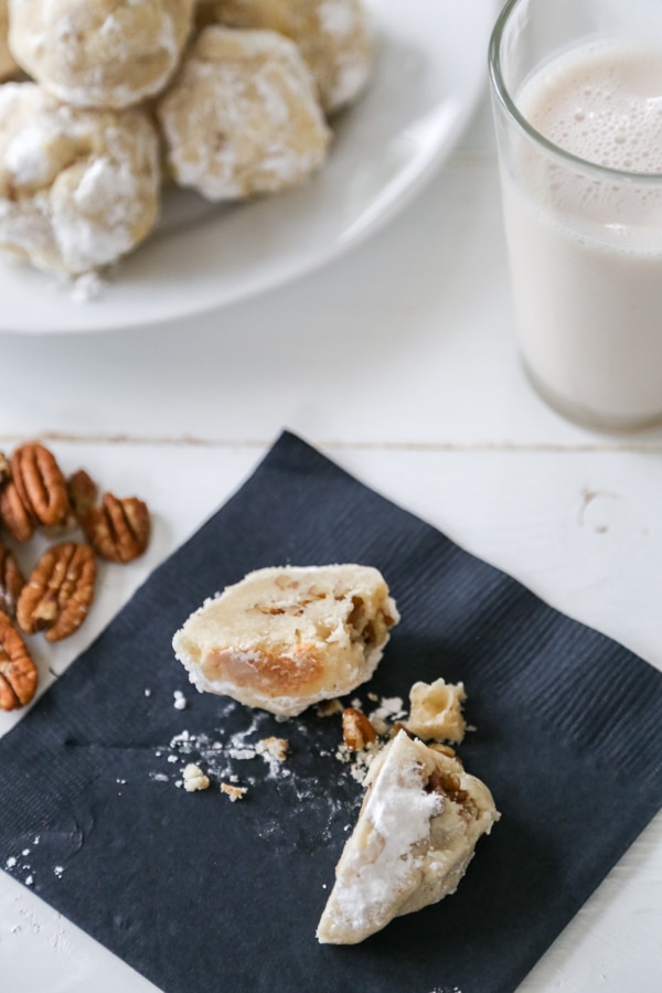A Russian Tea Cake broken in half on a black napkin with a plate of cookies and a glass of milk next to it. 