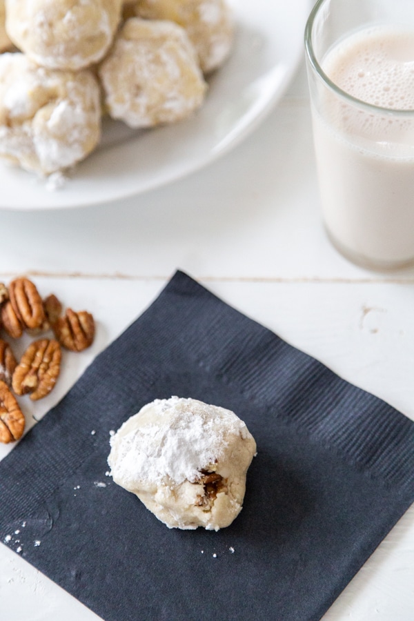 A black napkin with a Russian Tea Cake cookie and a white plate of cookies in the background. 