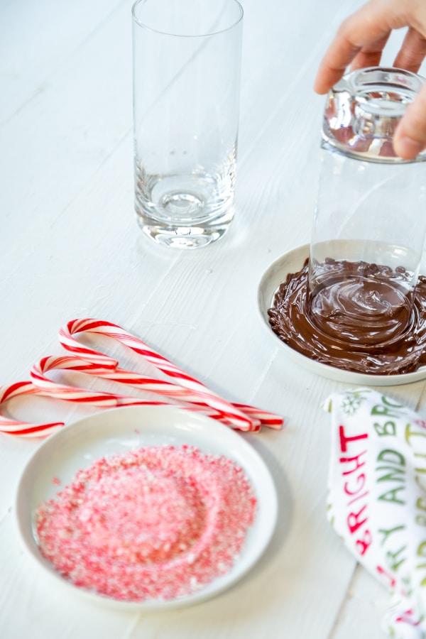 A hand dipping the rim of a glass in melted chocolate and a plate of ground candy cane.