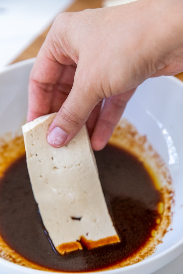 A hand dipping a strip of tofu into a bowl of marinade.